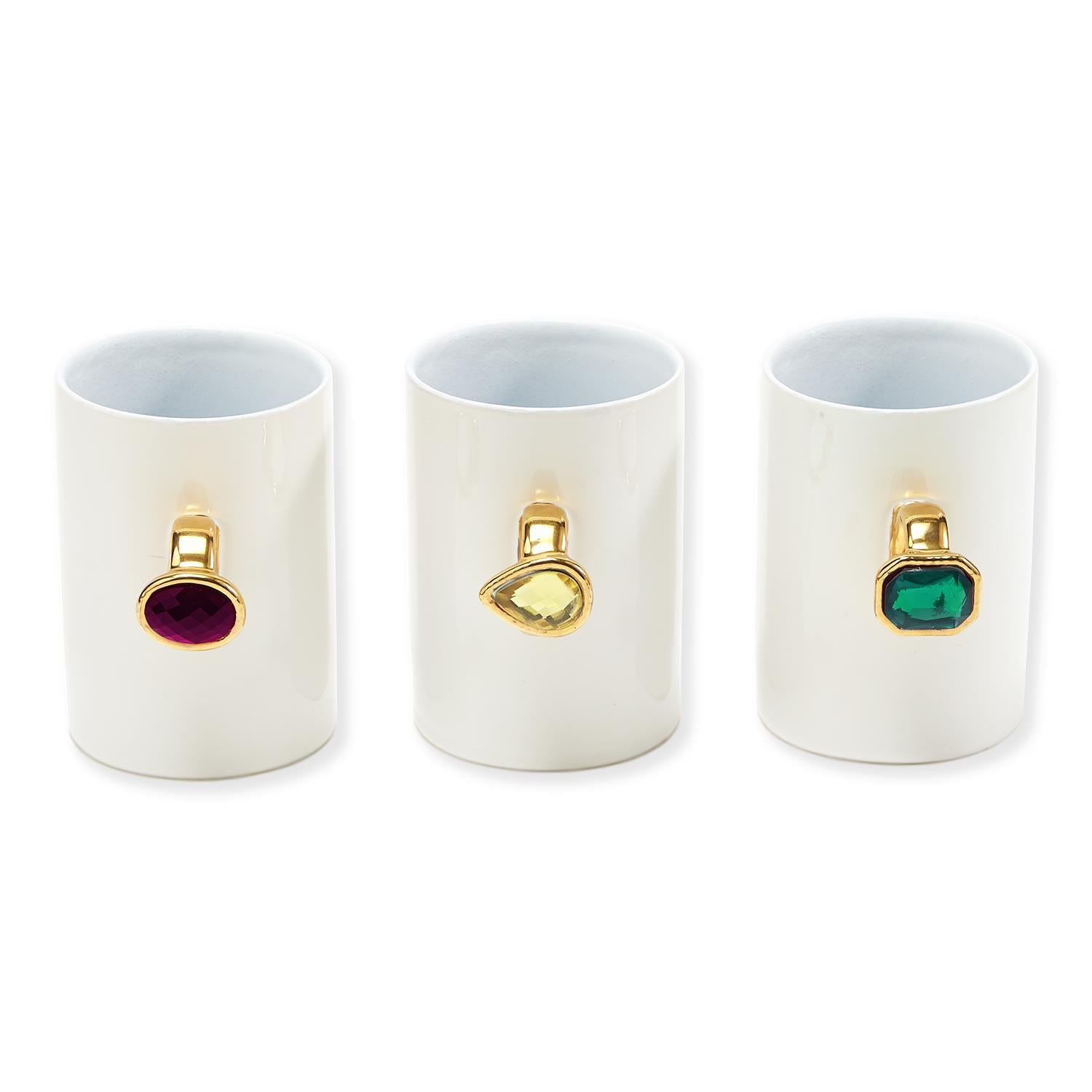 Three plain white mugs with gem rings for handles; the left mug has a pink oval gem ring, the middle mug has a yellow teardrop gem ring, and the right mug has a green emerald-cut gem ring.